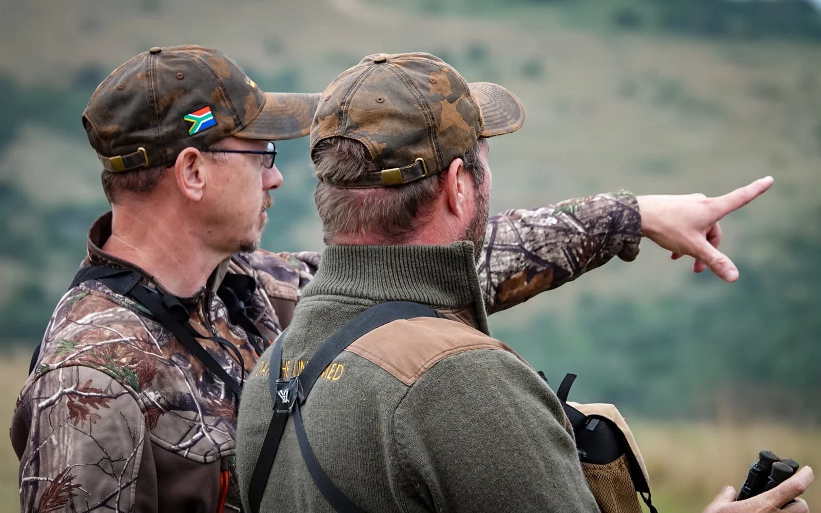 Two hunters looking out across a valley, one pointing into the distance