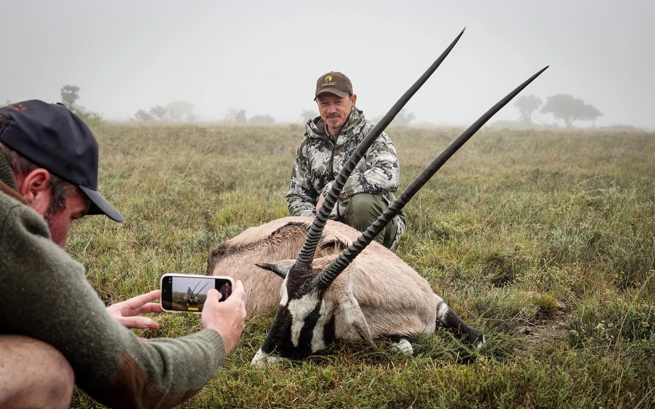 A hunter kneeling behind a horned antelope while a companion takes a photograph