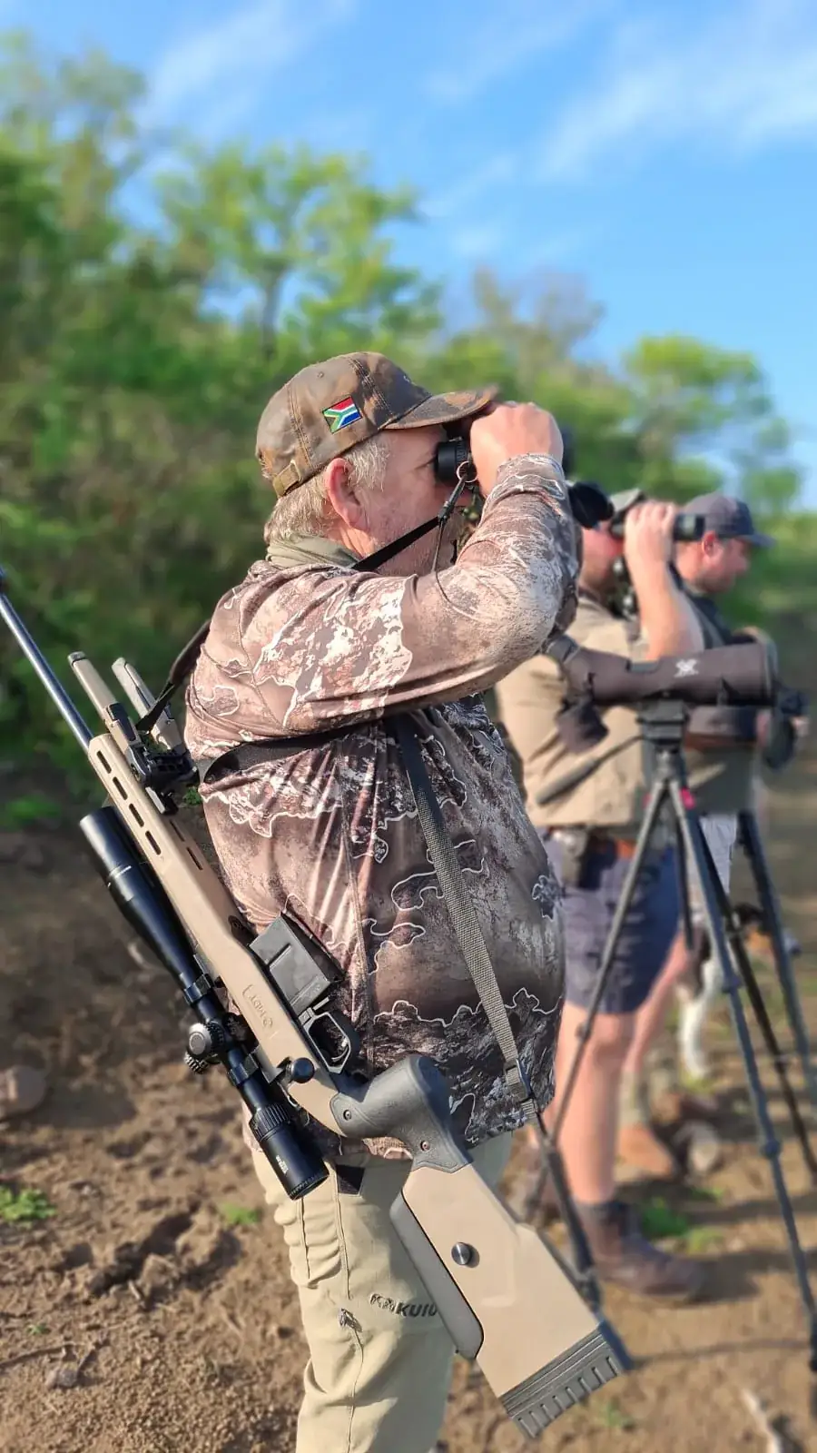Hunter looking through binoculars in the field with companions behind him